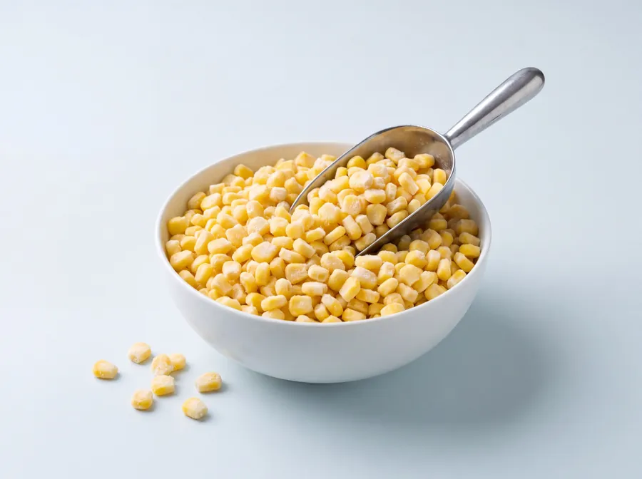 Frozen sweet corn kernels in a white bowl with a steel scoop